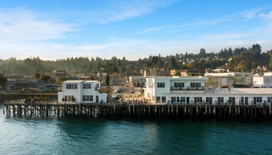 A coastal town with white buildings situated on a pier extends over calm water. The town is surrounded by forested hills under a clear blue sky. Small houses and buildings are visible in the background.