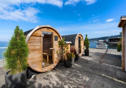 Two barrel-shaped wooden sauna cabins are situated outdoors near a waterfront. Potted plants are placed around the cabins, and a boat is visible in the water in the background. The sky is clear with a few clouds.