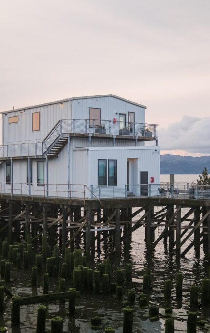 A waterside building on stilts is situated at the edge of a pier. The structure has two levels, with an exterior stairway leading to the upper level. The surrounding water is calm, and remnants of old wooden posts are visible in the foreground. Mountains and clouds appear in the background.