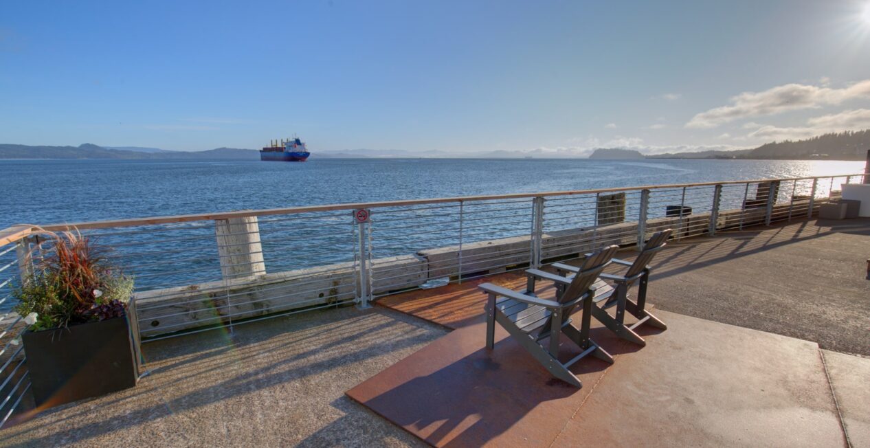 A waterfront scene with two lounge chairs facing the water on a sunny day. A cargo ship is visible in the distance. The area is bordered by a metal railing, and there are flowers in a planter box to the left. The sun is low in the sky, casting long shadows.