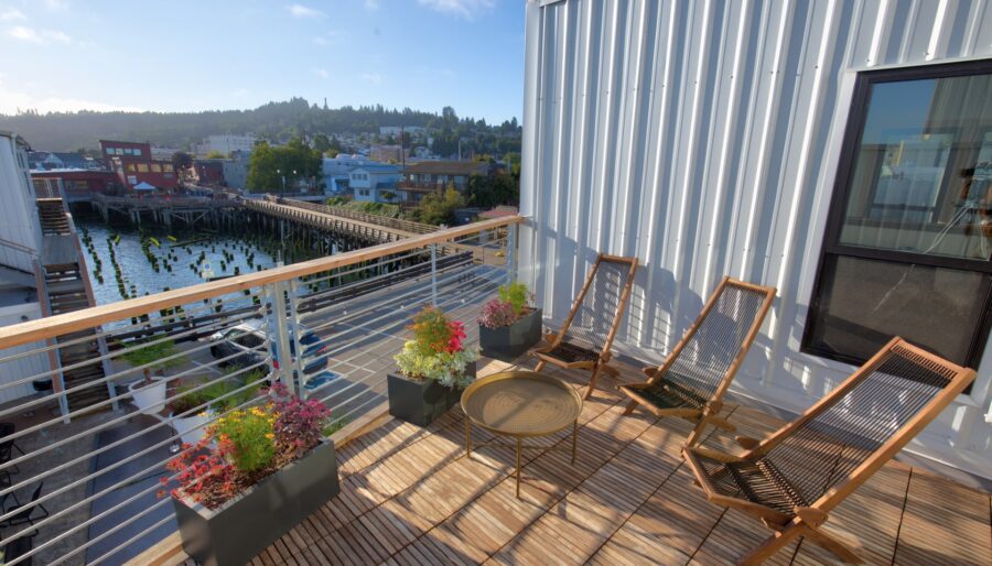 A wooden deck with three reclining chairs and a round table overlooks a coastal town. Potted plants line the railing, and wooden pilings are visible below near the water. The backdrop includes buildings and a hill with trees under a bright blue sky.