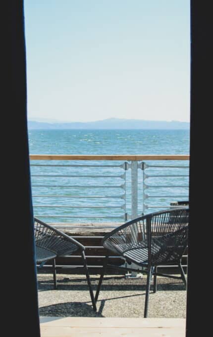 A view of the sea through a gap between two dark vertical panels. The scene includes two empty black wicker chairs facing a railing, with calm water and distant hills on the horizon under a clear sky.