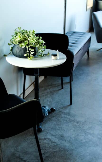 A small round white table with a black base is positioned between two black chairs. On the table, there is a black vase with green and white foliage and a small white container. A long gray padded bench is against the wall in the background.