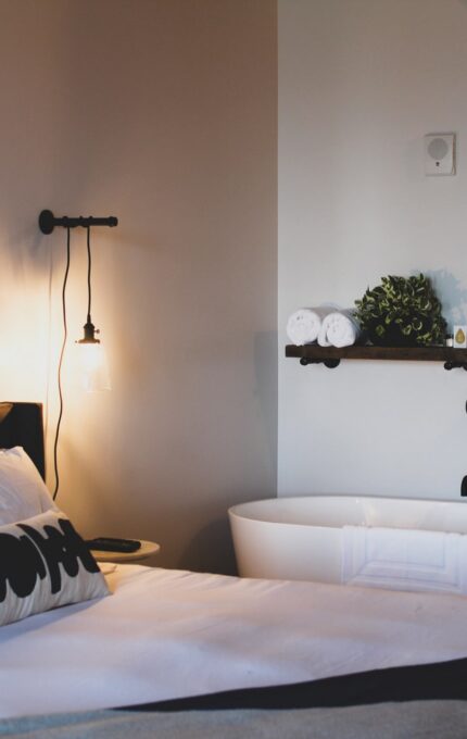 A bedroom features a bed with white linens, black and white pillows, and a round mirror on the wall above a bedside lamp. Adjacent to the bed is a white freestanding bathtub next to a rustic wooden column and a small shelf holding towels and greenery.