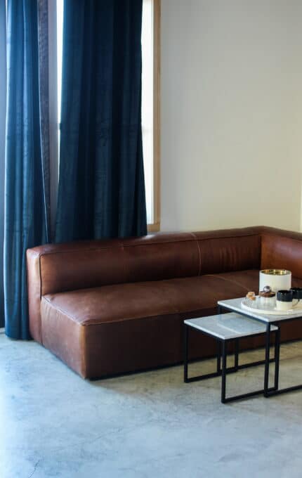 A modern living room corner features a brown leather sectional sofa, a light-colored floor, a dark curtain, and a set of two small square coffee tables with metal legs. Various decorative items are placed on the tables. Green plants are partially visible.