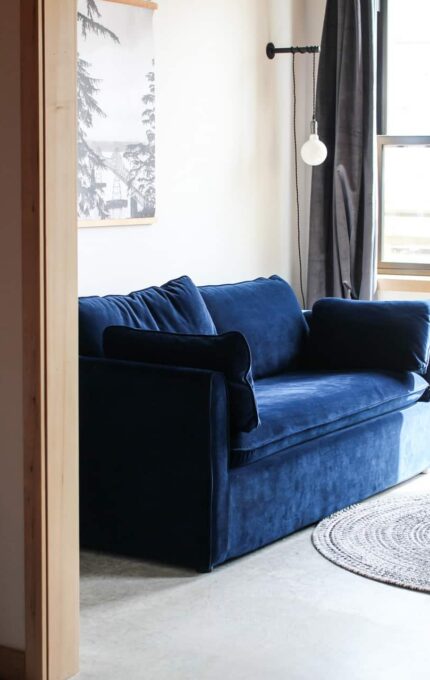 A room with a blue sofa against the wall, positioned next to a window with curtains. A round, woven rug sits on the floor in front of the sofa. A small, round coffee table is placed on the rug. The room also has a framed picture on the wall.