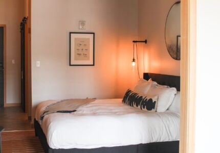 A cozy bedroom with a neatly made bed featuring white linens and several decorative pillows. Above the bed, a round mirror and framed artwork are mounted on the wall. A pendant light hangs from the wall on the right side of the bed.