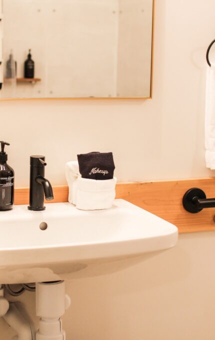 A small bathroom with a white sink, a black faucet, and a black soap dispenser on the left. A wall-mounted mirror is above the sink. A white folded towel and a black hand towel are on the right, along with a black towel ring and a grab bar.