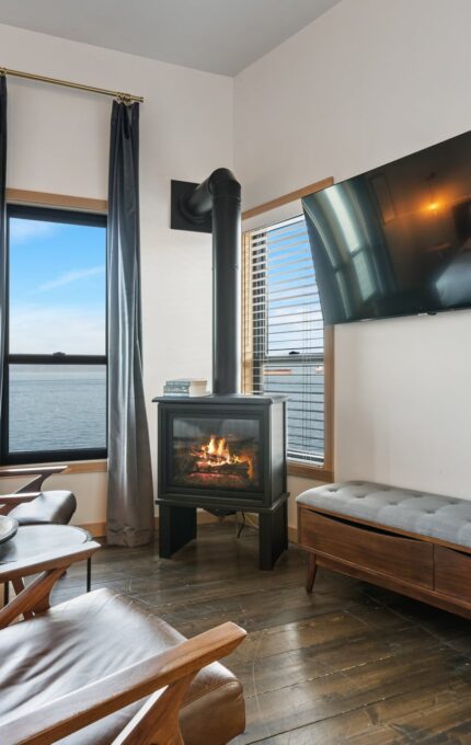 A modern living room features two brown leather armchairs facing a fireplace and a wall-mounted flat-screen TV. Large windows offer a scenic view of the water. A wooden bench sits below the TV, and a round table with plates and a teacup is between the chairs.