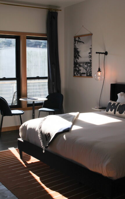 A sunlit bedroom with a large bed featuring white bedding and a gray throw blanket. Beside the bed is a wall-mounted lamp and a "Home" pillow. Near the window are two chairs and a small table. The room has a modern design, with a framed photo hanging on the wall.