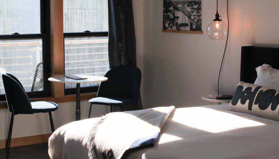 A sunlit bedroom with a large bed featuring white bedding and a gray throw blanket. Beside the bed is a wall-mounted lamp and a "Home" pillow. Near the window are two chairs and a small table. The room has a modern design, with a framed photo hanging on the wall.