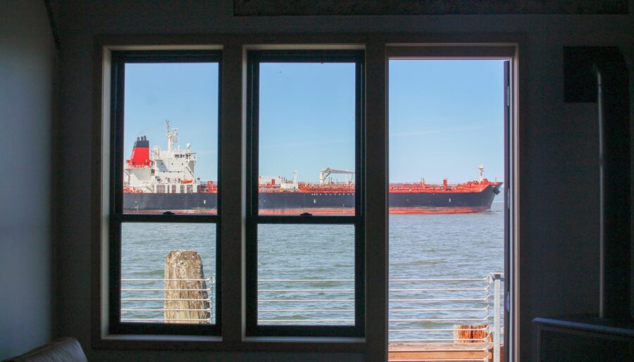 A large cargo ship is visible through two windows and a door of a room overlooking the water. The ship is painted red and black and is moving from left to right.