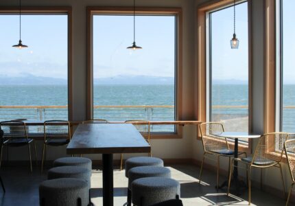 A bright, modern café with large windows overlooking a calm ocean view. The seating includes bar stools along the windows, a long wooden table with stools on either side, and a small table with chairs by the window. Pendant lights hang from the ceiling.