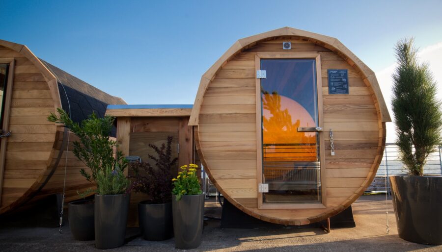 A rounded wooden sauna sits on a concrete surface with a glass door reflecting an orange sunset. There are several potted plants arranged in front of the sauna, and a metal railing is visible in the background with a view of the sea and sky.