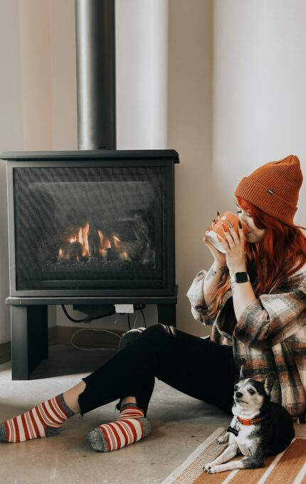 A person with long red hair, wearing a red beanie, plaid shirt, and striped socks, sits on the floor near a lit fireplace. They are holding a mug and there is a small dog sitting beside them. The person appears to be enjoying their drink.