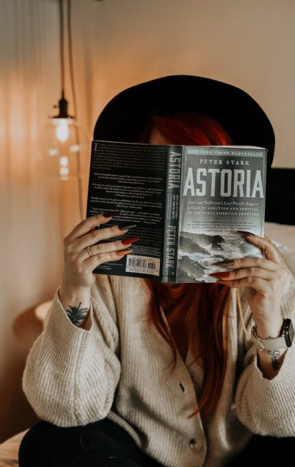 A person with long red hair, wearing a black hat and beige cardigan, reads a book titled "Astoria" by Peter Stark. They are sitting in a cozy, well-lit room with a hanging light bulb in the background. Their face is obscured by the book.