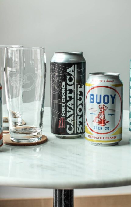 A round white marble table displays four different beer cans and three empty beer glasses. The beer cans include Buoy Beer Co. Cream Ale, Fort George Cavatica Stout, Buoy Beer Co. Czech-Style Pilsner, and Gray IPA.