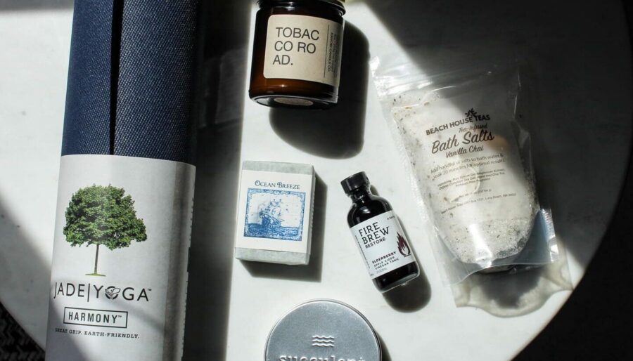 A flat lay of wellness items on a white surface, including a rolled navy yoga mat, a candle, a bar of soap, a jar of bath salts, a small bottle, and a round tin.