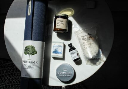 A yoga mat, candle, soap, bath salts, essential oil, and shaving balm are arranged on a circular white table. The yoga mat is dark blue. The candle reads "Tobacco Road." The bath salts are labeled "French White Natural Bath Salts.