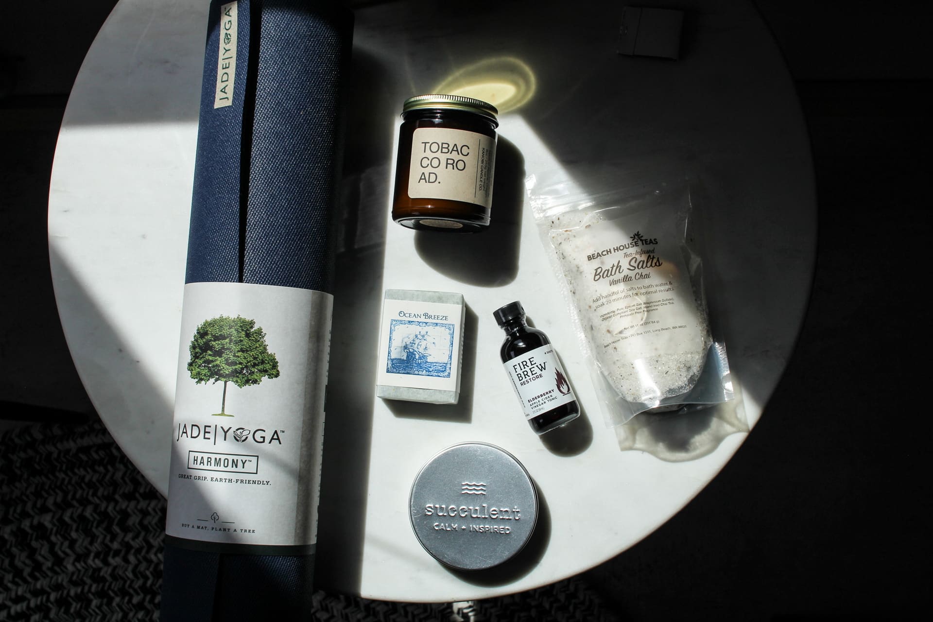 A yoga mat, candle, soap, bath salts, essential oil, and shaving balm are arranged on a circular white table. The yoga mat is dark blue. The candle reads "Tobacco Road." The bath salts are labeled "French White Natural Bath Salts.