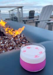 A pink cocktail with foam and heart-shaped designs on top sits on a table beside a gas fire pit with glowing flames. In the background, there are Adirondack chairs overlooking a cloudy sky and a distant view of water.