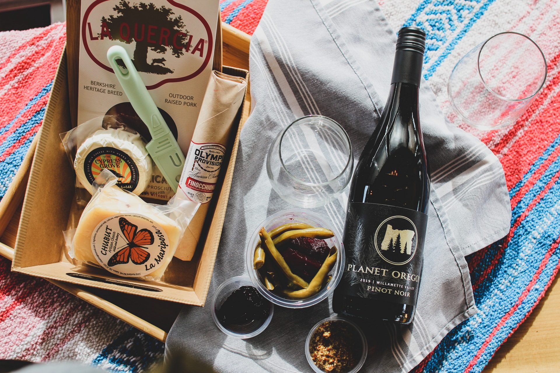 A box containing various gourmet food items like La Quercia Berkshire Prosciutto, a round of aged cheese, mustard, and green beans in a jar. Next to the box is a bottle of Planet Oregon Pinot Noir and three empty glasses on a striped cloth.