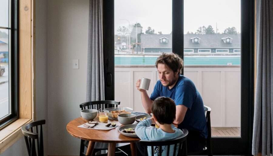 An adult and a child sit at a round wooden dining table set with plates and bowls, eating a meal together in a bright room with large windows and glass doors. The adult holds a white mug.
