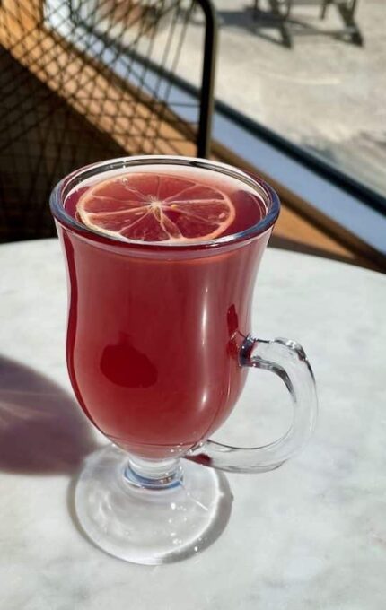 A clear glass mug, filled with a red liquid and garnished with a slice of lemon, is placed on a round, white marble table. The background shows a sunny outdoor setting with a window and a wire-frame chair partially visible.