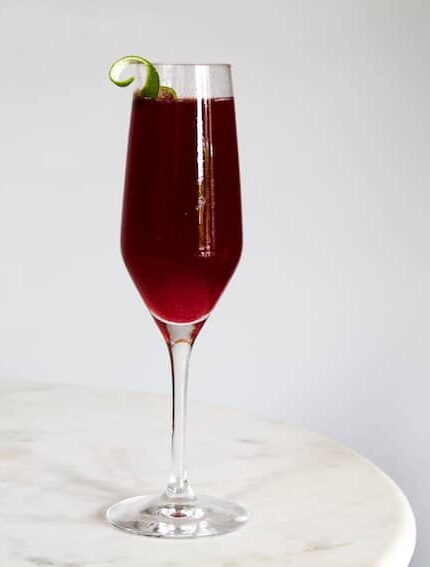 A tall champagne flute filled with a deep red beverage sits on a white marble surface. The drink is garnished with a small, twisted lime peel on the rim of the glass. The background is a plain, light gray wall.