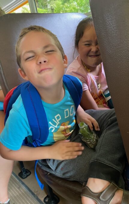 Two children are sitting on a bus seat. The child on the left is wearing a blue shirt and a blue backpack, making a playful face. The child on the right, dressed in a pink shirt, is also smiling. Both appear to be enjoying the bus ride.