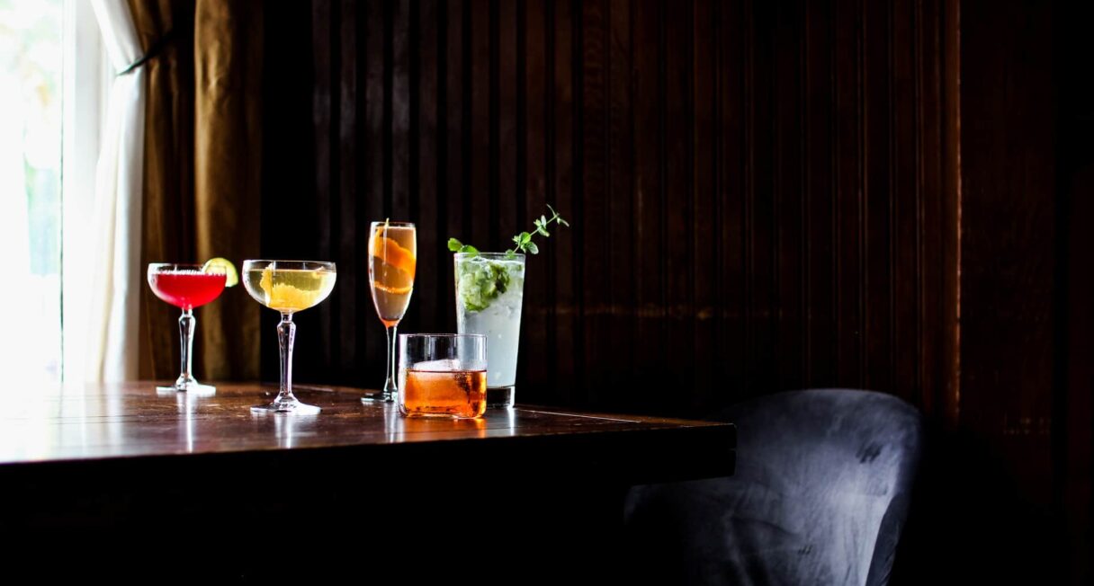 A wooden table holds five cocktails in various glasses, featuring vibrant colors like red, gold, and green. The background is dark with wooden paneling, and a soft light filters through a window with a curtain. A dark chair is partially visible at the edge of the table.