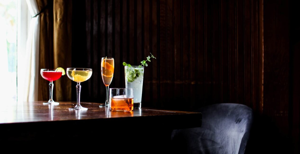 A wooden table holds five cocktails in various glasses, featuring vibrant colors like red, gold, and green. The background is dark with wooden paneling, and a soft light filters through a window with a curtain. A dark chair is partially visible at the edge of the table.