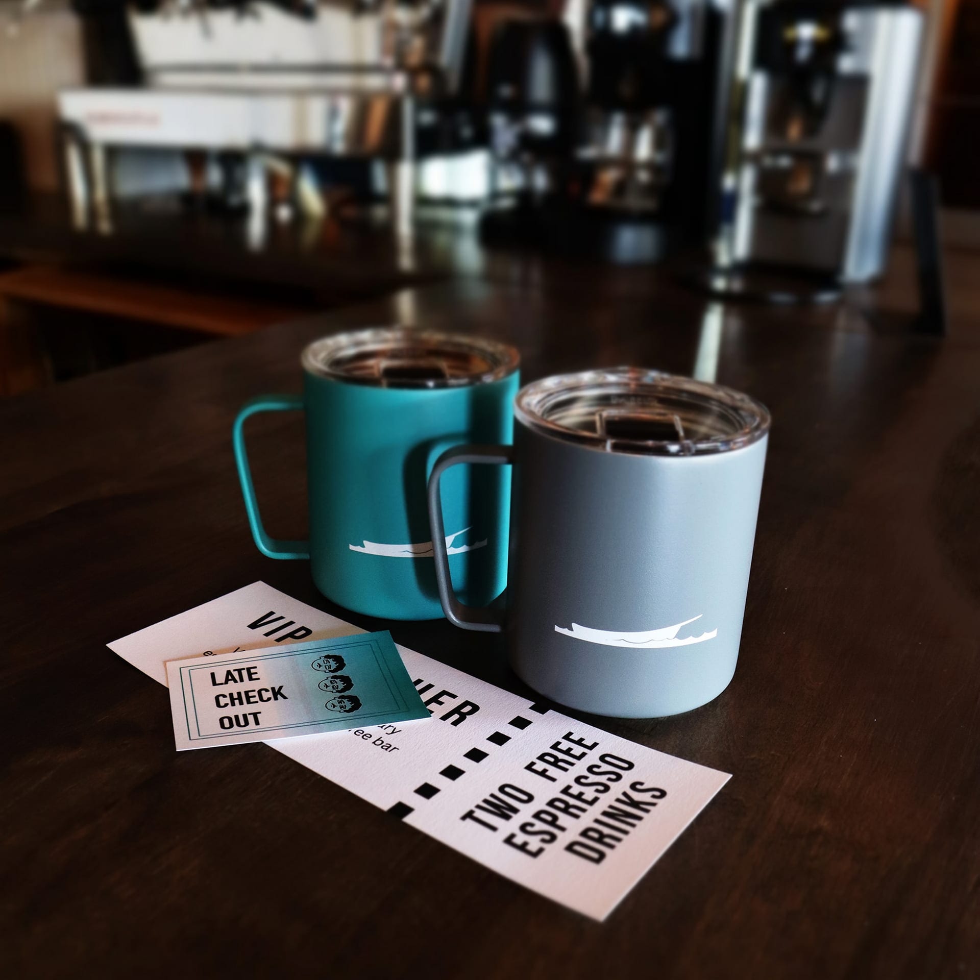 Two insulated mugs, one teal and one gray, sit on a dark wooden table. Next to them are two cards: one offering two free espresso drinks and the other a late checkout. A coffee machine is blurred in the background.