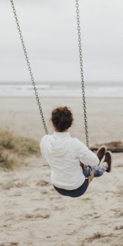 Two people are sitting on swings facing the ocean at a beach on an overcast day. The person on the left is wearing a white hoodie, and the person on the right is wearing a gray jacket and a red scarf. The beach is mostly empty with a few people in the background.