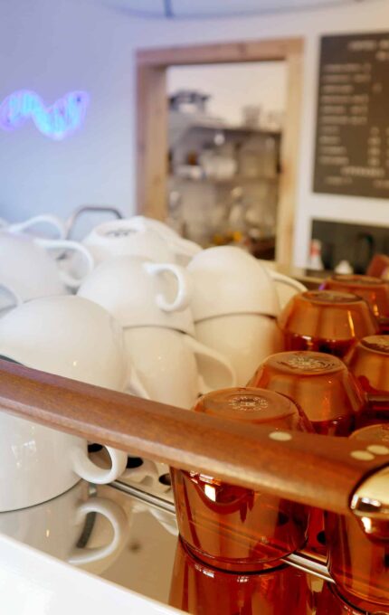 A coffee shop counter with a display of stacked coffee cups, white on the left and amber on the right. A menu board with blurred text is visible in the background, along with a neon sign and various equipment.