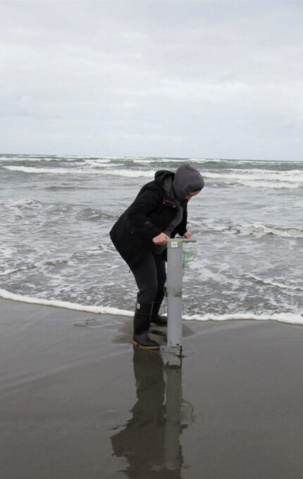 A person standing on a beach, wearing a hooded jacket and boots, is operating a tall cylindrical device in the wet sand near the shoreline. The sky is overcast, and waves are gently rolling onto the shore.