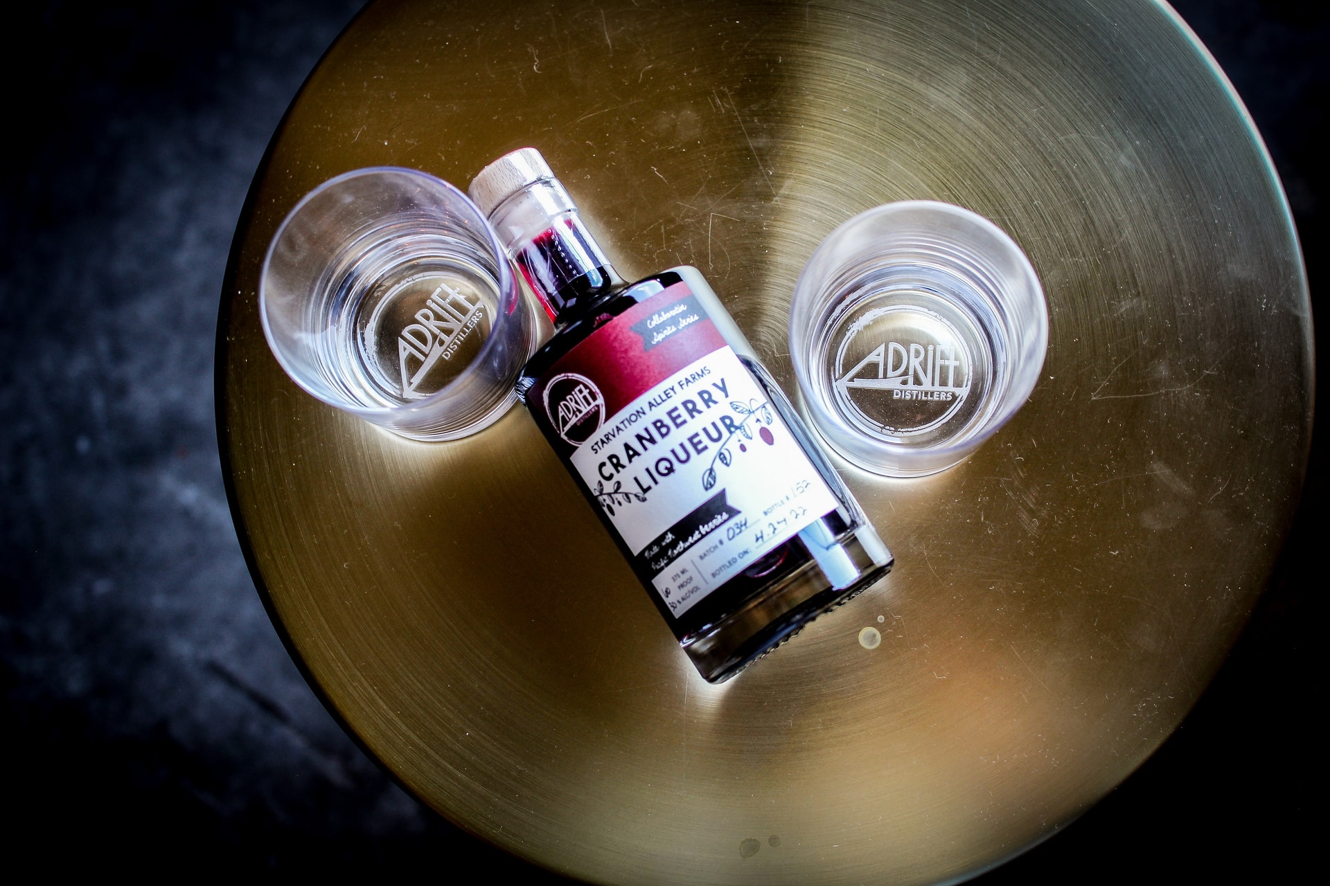 A bottle of cranberry liqueur is placed on a round, golden table, accompanied by two clear glasses that have "Adria" printed on them. The bottle lays horizontally in between the two upright glasses.