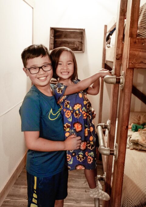 A young boy wearing glasses and a blue outfit is holding a smiling girl in a floral dress as she climbs a wooden bunk bed ladder. The bunk bed is located in a brightly lit room.