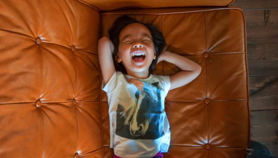 A child lays back on a brown tufted leather sofa, laughing with eyes closed and arms behind their head, wearing a t-shirt with a horse graphic and pink pants. The photo is taken from above.