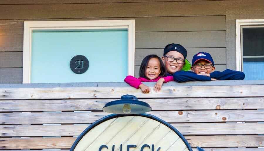 Three smiling children lean over a wooden balcony railing above a large round sign that reads “CHECK IN eat. drink.” A door with the number 21 is visible in the background.