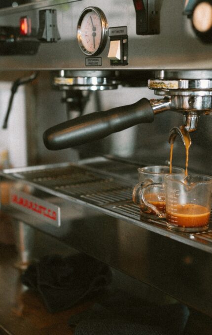 A close-up of an espresso machine making two shots of espresso. The streams of coffee are being dispensed into two small glass cups placed on the machine's drip tray. The espresso machine has various gauges and knobs, with some other items in the background.