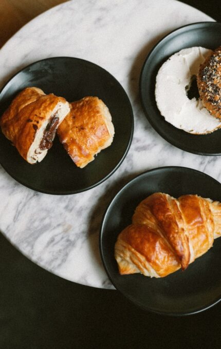A round marble table with three black plates. One plate has a chocolate croissant, another has a bagel with cream cheese and seeds, and the third plate has a plain croissant. Part of a wooden surface is visible on the left.