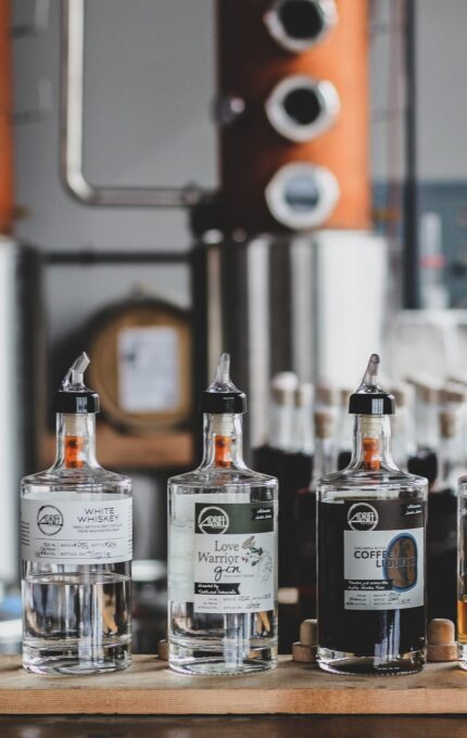 A line of various bottled spirits is displayed on a wooden surface in front of distilling equipment. The bottles are labeled with different types of alcohol, including vodka, gin, coffee liqueur, and cranberry liqueur, among others.