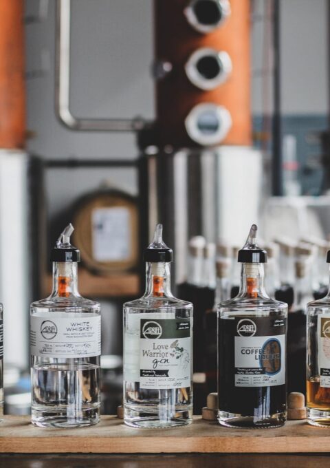 A line of various bottled spirits is displayed on a wooden surface in front of distilling equipment. The bottles are labeled with different types of alcohol, including vodka, gin, coffee liqueur, and cranberry liqueur, among others.