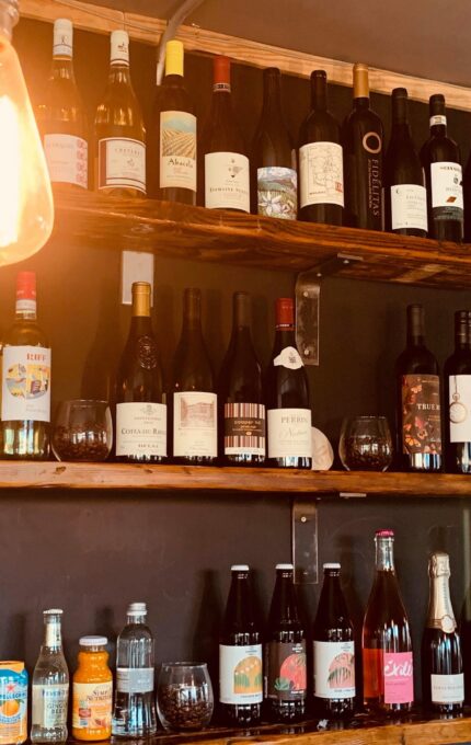 Shelves displaying an assortment of bottles, primarily wine with labels visible on the upper shelves. Below, a variety of other beverages, including soft drinks and water, are arranged. A glowing light bulb hangs in the upper left corner.