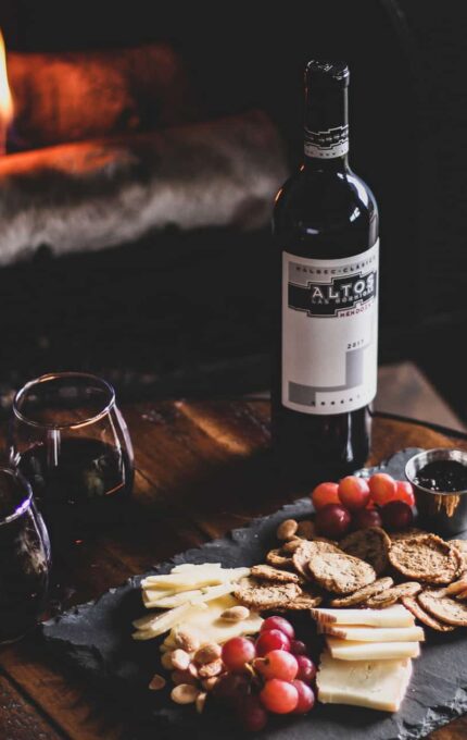 A bottle of wine, two glasses with red wine, and a charcuterie board with assorted cheeses, crackers, nuts, grapes, and a small container of jam are on a round wooden table in front of a lit fireplace.