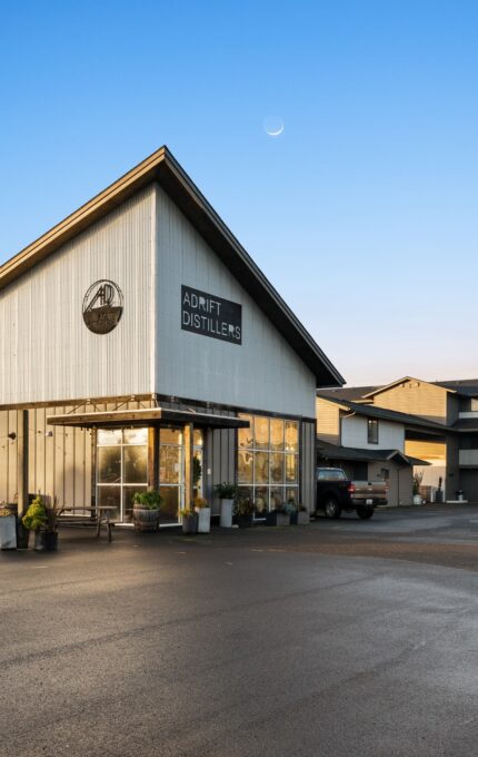 A modern white building with a triangular roof that houses the "Journey Distillery." The entrance is glass-walled, and a black truck is parked in front. Another multi-story building is visible in the background. The sky is clear with a crescent moon.