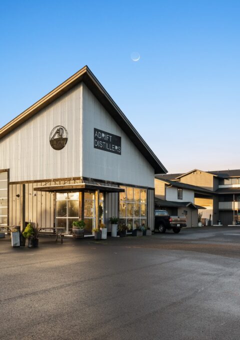 A modern white building with a triangular roof that houses the "Journey Distillery." The entrance is glass-walled, and a black truck is parked in front. Another multi-story building is visible in the background. The sky is clear with a crescent moon.