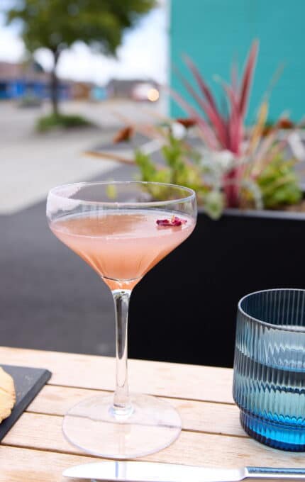 A close-up of a pink cocktail in a glass on an outdoor table. A knife rests on the table beside the glass. In the background, there's a potted plant and a blurry exterior view with a tree and a teal-colored wall.