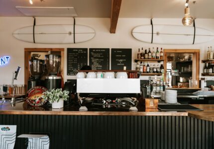 A coffee shop interior with a wooden counter, espresso machine, and coffee grinder. Shelves on the back wall display various bottles and coffee equipment. Two surfboards are mounted on the wall above the counter. A neon "OPEN" sign is to the left.